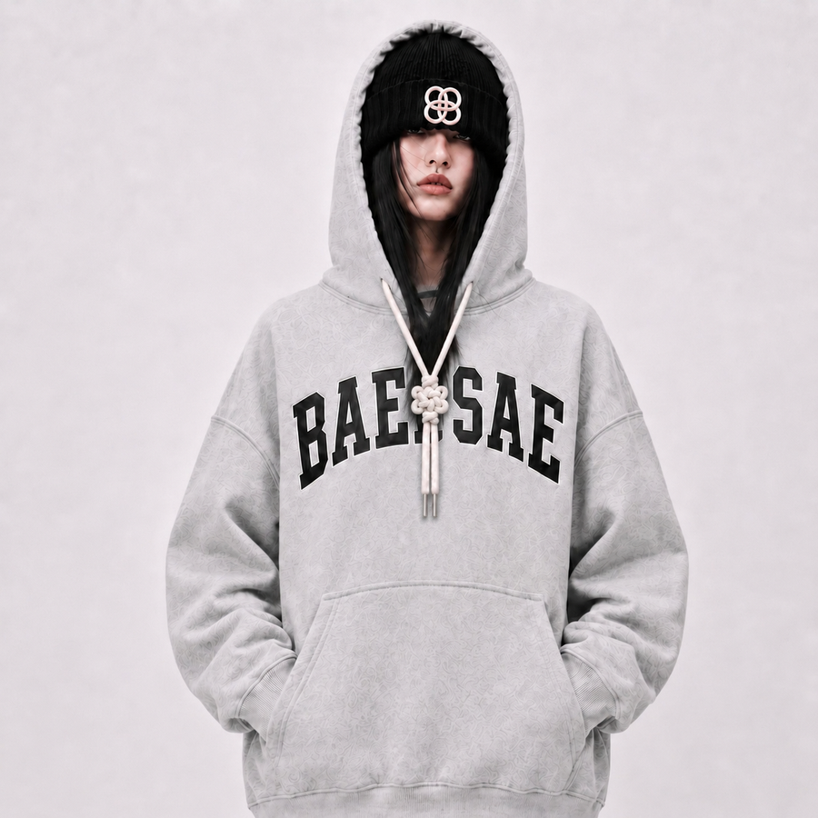 Person wearing a gray hoodie with 'BAEWSAE' text, black beanie, and long necklace on a light gray background